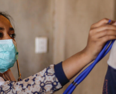 A woman doctor, masked, listening to a child's chest through a stethoscope. Image credit: Save the Children