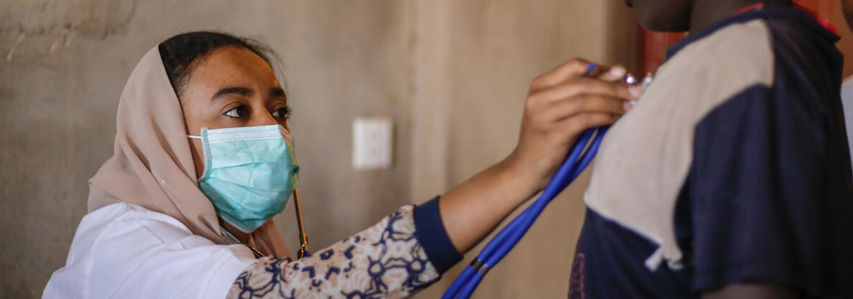 A woman doctor, masked, listening to a child's chest through a stethoscope. Image credit: Save the Children